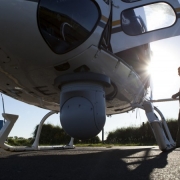 Visa da parte inferior frontal de um helicóptero da brigada militar, equipado com câmeras. Ao lado um homem caminha em direção ao helicóptero.