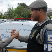 Brigadiana cumprimentando com a mão direita outro brigadiano, em frente a uma viatura com a inscrição "Patrulha Maria da Penha"