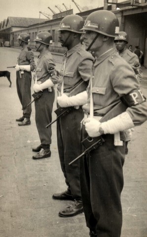 Quatro homens parados, de uniforme e capacete, segurando armas viradas para baixo em foto tirada de ângulo lateral. 