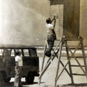 um homem parado ao lado de uma van estacionada, observando um segundo homem que está subindo em uma cabine de salva vidas. Os dois de camiseta clara, na praia, com mar ao fundo.