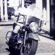 Homem uniformizado de cap, sorrindo para a foto em cima de uma moto.