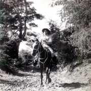 Homem uniformizado, de chapéu com abas, andando a cavalo em um campo com arvores.