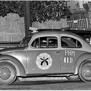 Antiga viatura da PRM estacionada na rua, em frente ao muro de uma casa, com um homem passando atrás do carro.