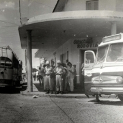 rua com dois ônibus estacionados em frente a prédio, com grupo de oficiais da brigada embaixo do teto da varanda, na sombra.