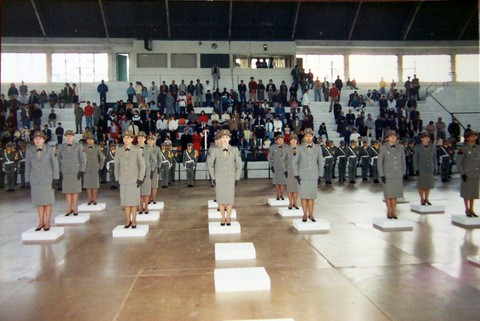 Grupo de brigadianas do sexo feminino, agrupadas em seis filas, uma do lado da outra em um ginásio. Ao fundo um grupo de pessoas em uma arquibancada.