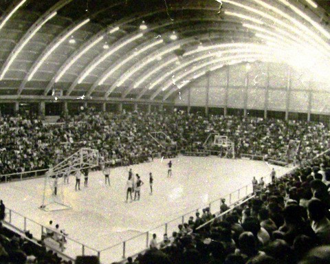 Estádio lotado de pessoas assistindo a um jogo de basquete, com grupo de jogadores no meio da quadra.