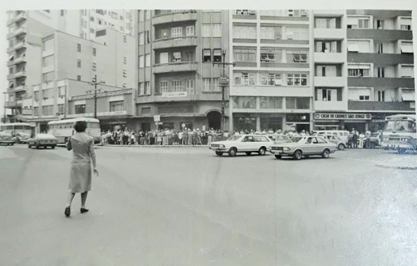 Rua com prédios ao fundo, carros andando, uma mulher atravessando a rua e multidão de pessoas ao fundo.