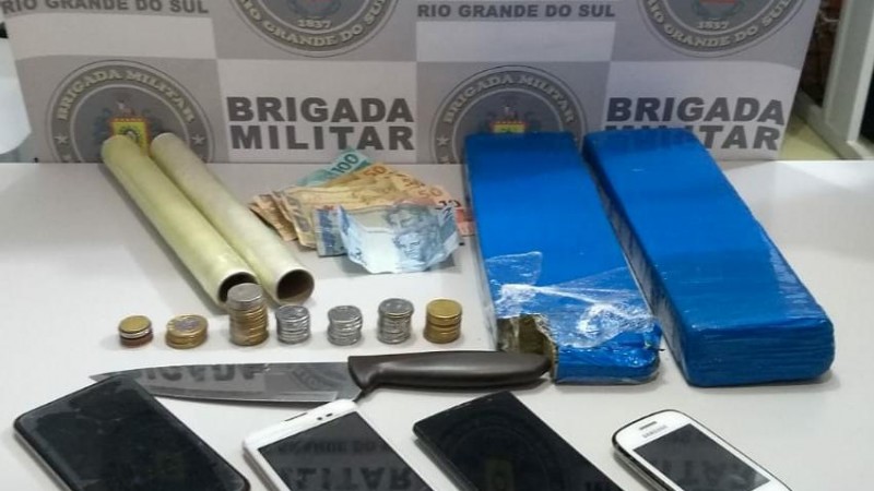 Materiais apreendidos como dorgas, faca, celulares, moedas, dinheiro com o banner da Brigada Militar ao fundo