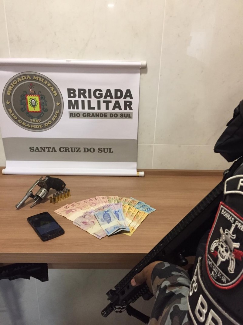 Revólver, celular e cédulas de dinheiro no fundo um banner da Brigada Militar