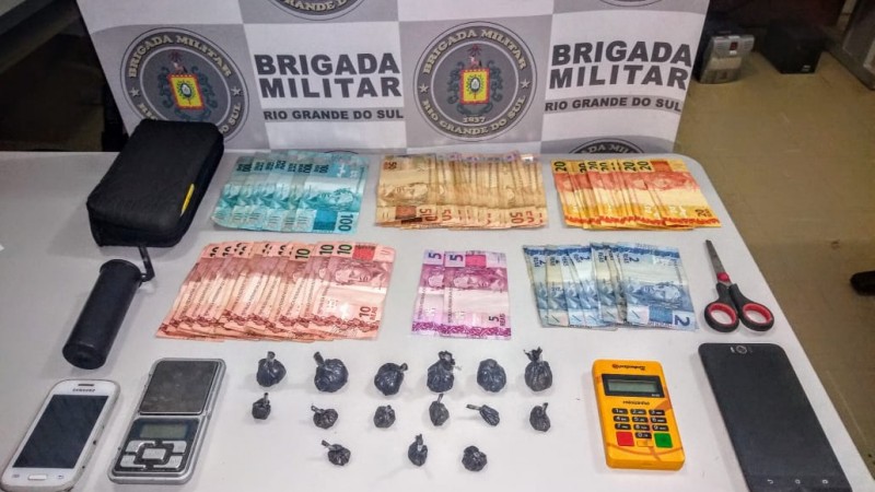 Uma mesa com os materiais apreendidos (Drogas, dinheiro, celulares, tesoura, entre outros) e no fundo o banner da Brigada Militar