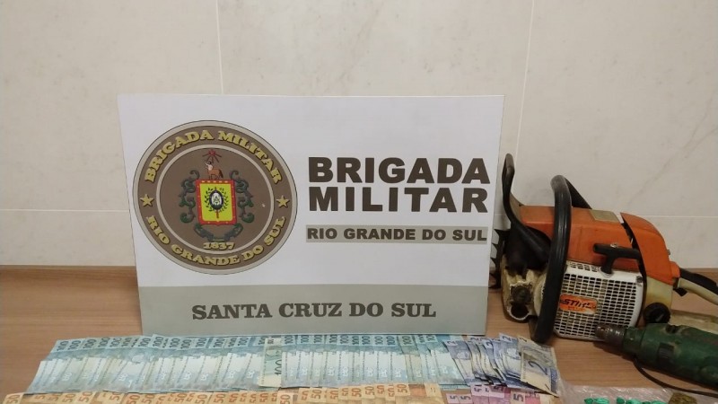 Uma mesa contendo nela o material apreendido (Dinheiro, motoserra, furadeira, drogas fracionadas) e no fundo o banner da Brigada Militar.
