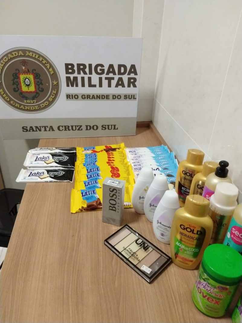 Uma mesa com o fundo o banner da Brigada Militar. Sobre a mesa esta o material apreendido (Chocolates, shampos, material de higiene e maquiagem)