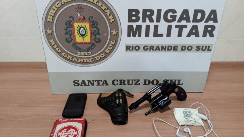 Uma mesa com os materiais apreendidos (arma, telefone, fone de ouvido) e no fundo o banner da Brigada Militar