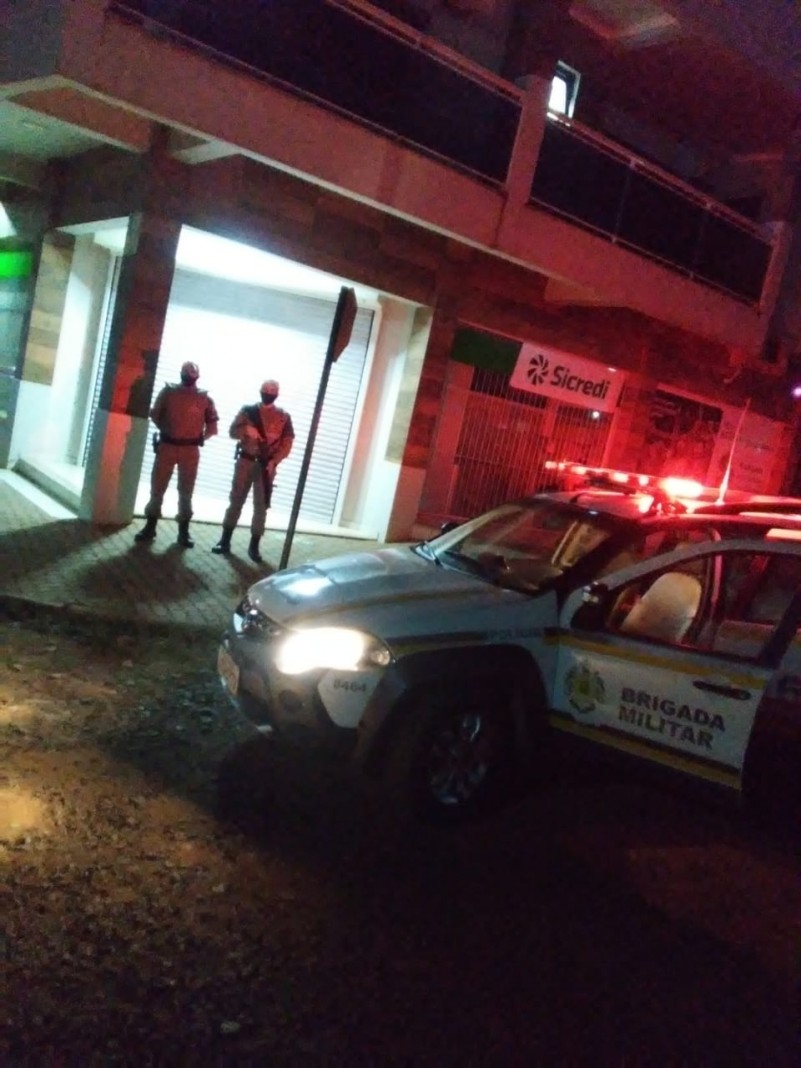 Uma viatura da Brigada Militar a frente e dois policiais militares ao fundo fazendo policiamento ostensivo em frente a uma agência bancária