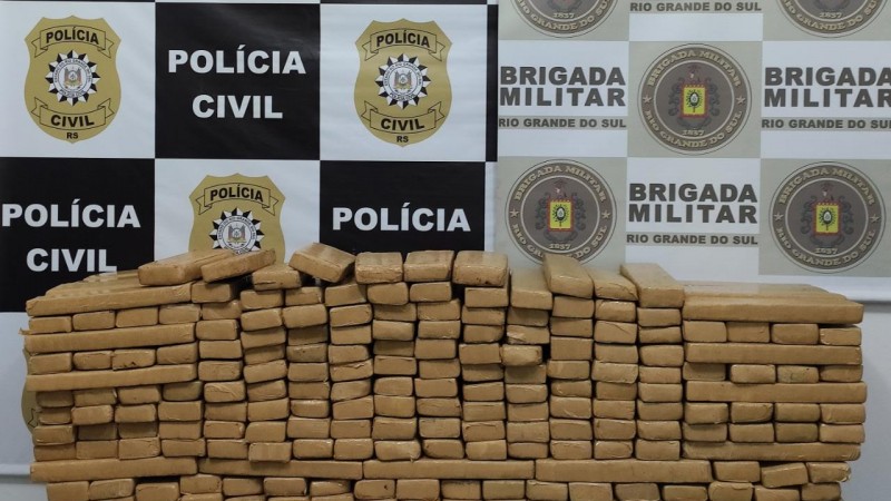 Na imagem uma pilha de tijolos de drogas embaladas. Ao fundo dois banners, um com s&iacute;mbolos da Policia Civil e outro com s&iacute;mbolos da Brigada Militar