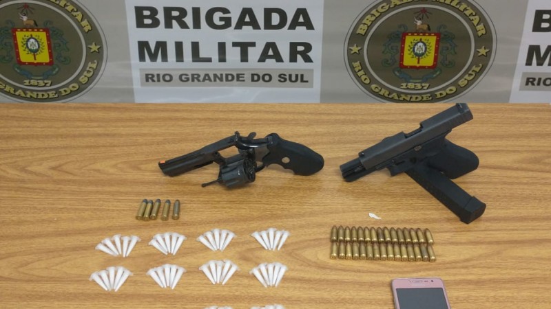 Na foto est&atilde;o dispostas em uma mesa as armas, muni&ccedil;&otilde;es, drogas e dinheiro apreendidos. No fundo o banner da Brigada Militar