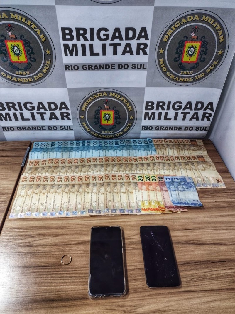 Banner da Brigada Militar ao fundo. Sobre uma mesa dinheiro em esp&eacute;cie, celulares e uma alian&ccedil;a.
