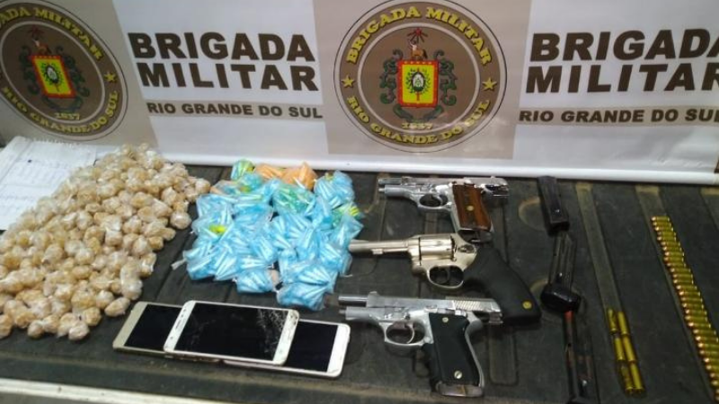 Na foto diversos pacotes e pinos de drogas, armas, munições e três celulares. Ao fundo o banner da Brigada Militar