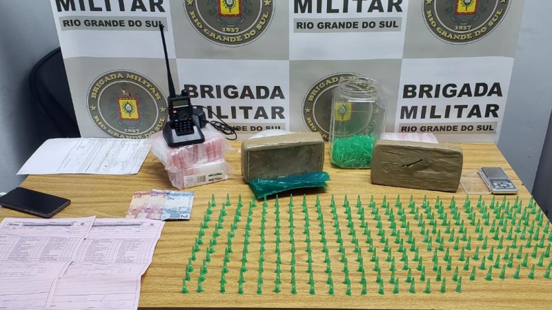 Ao fundo da imagem o banner da Brigada Militar.  Na frente do banner est&aacute; uma mesa onde todos os itens apreendidos est&atilde;o expostos. Dentre esses itens est&atilde;o tijolos de drogas, pinos de drogas e outros materiais