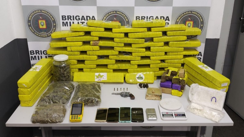 A imagem mostra tijolos de maconha, arma, munições  entre outras drogas sobre uma mesa. Ao fundo o banner da Brigada Militar.