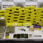 A imagem mostra tijolos de maconha, arma, munições  entre outras drogas sobre uma mesa. Ao fundo o banner da Brigada Militar.