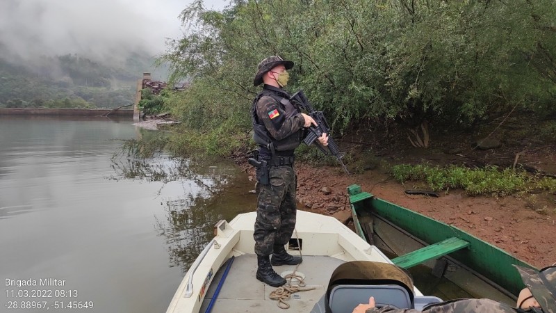 Policial Militar, em pé em um barco pequeno, segura um fuzil, às margens de um Lago pequeno, onde está localizada a Pequena Central Hidrelétrica, na cidade de Vila Flores. O tempo está nublado e aparecem árvores e vegetação verde às margens do Lago.