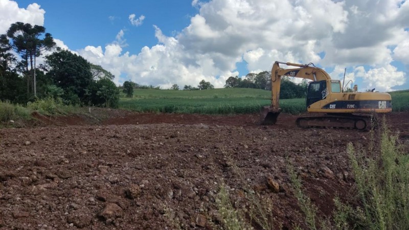 Área de terra sendo manipulada por uma máquina retroescavadeira, de cor amarela, da marca CAT. Ao fundo um área verde com árvores e um campo. O céu está azul com muitas nuvens brancas.