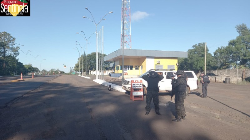 Dois Policiais Militares, vestindo fardas camufladas em tons de verde, ao lado de dois agentes da Secretaria da Agricultura, vestindo roupas escuras, estão às margens de uma rodovia.  Uma caminhoneta de cor branca encontra-se estacionada ao lado dos Policiais.  Ao fundo, um prédio pequeno, de um andar , de cor amarela e telhado azul claro. Uma antena de transmissão está posicionada atrás do prédio. Há árvores emoldurando a foto. O céu está limpo e azul.