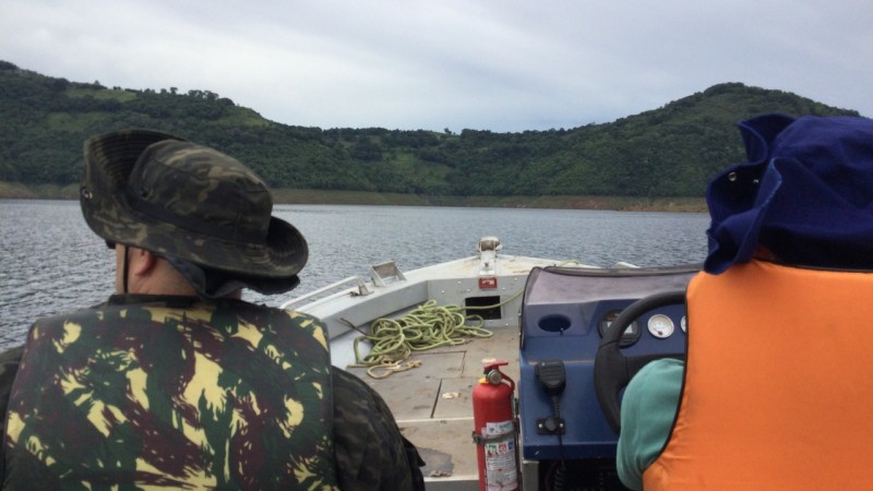 Patrulhamento Embarcado em Pinhal da Serra - Na foto, um Policial fardado com fardamento camuflado está ao lado de um Técnico do meio ambiente que está vestido de azul e usa um colete salva-vidas de cor laranja. os homens estão de costas, dentro de um barco pequeno. Ao fundo aparece um lago com águas escuras e uma Mata fechada