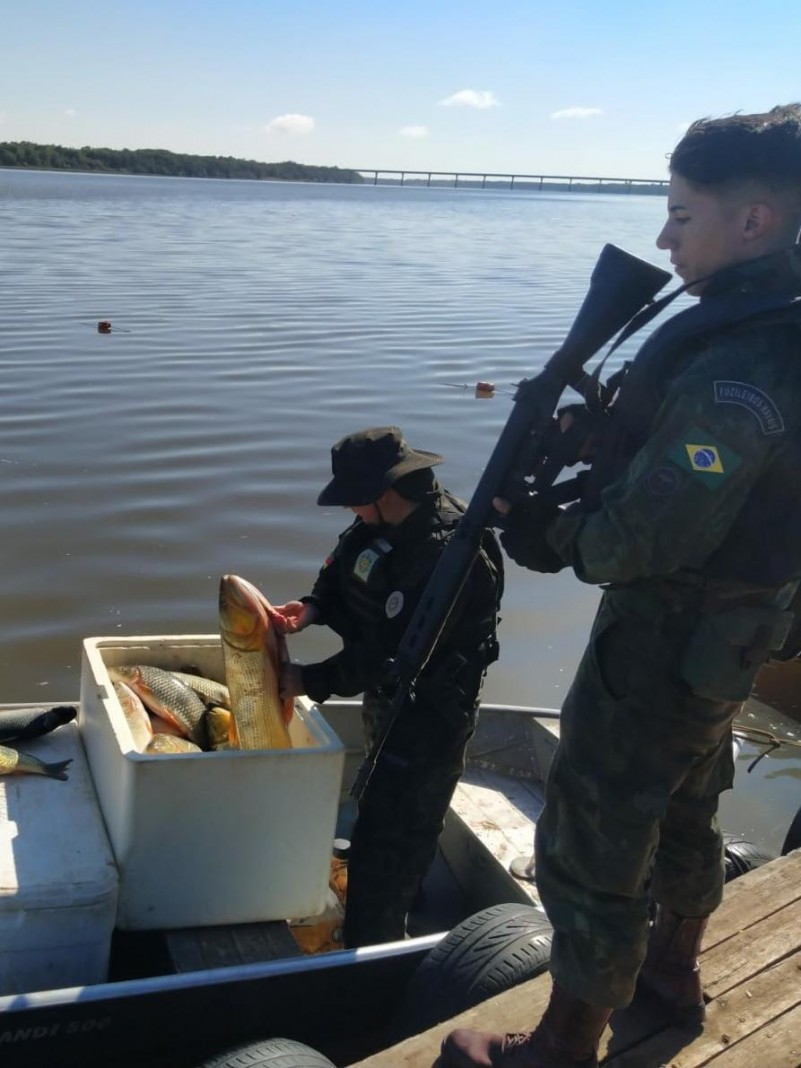 Um Fuzileiro Naval, com fardamento camuflado, segura um fuzil, ao lado de um barco pequeno atracado em um Cais. No barco um Policial Militar fardado com farda camuflada, segura um peixe da espécie do Dourado na mão direita e fiscaliza uma caixa de isopor branca contendo muitos peixes. Ao fundo o Rio Uruguai com águas escuras. O céu está azul e sem nuvens.