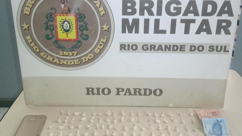 Material apreendido a frente e ao fundo o banner institucional da Brigada Militar de Rio Pardo.
