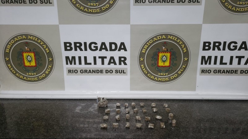 Drogas expostas em mesa com o banner da Brigada Militar ao fundo.