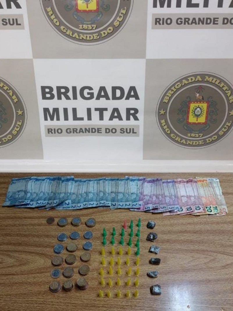 Drogas apreendidas em prisão por tráfico disposta à mesa com o banner da Brigada Militar ao fundo