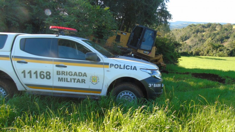 Foto Colorida. No primeiro plano, uma viatura policial, branca, com listras amarela e verde militar, com o brasão da Brigada Militar, parada ao lado de um trator de cor amarela. Ao fundo, uma mata verde, com campo verde ao redor. Há sol .