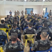 Grupo de homens fardados sentados em cadeiras azuis e amarelas em uma sala branca de costas para a câmera. A frente deles estão militares de pé lado a lado.