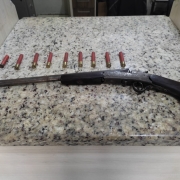 Foto mostra uma espingarda em cima de uma mesa, junto a munições.