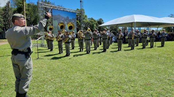 Imagem mostra banda de música da BM tocando em campo aberto durante uma formatura da corporação.