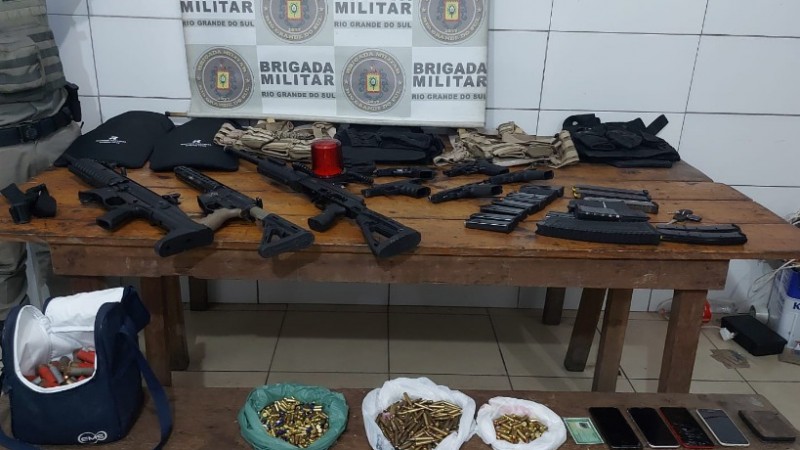 Imagem mostra uma mesa de madeira com muitas armas em cima. Por trás, o banner da Brigada Militar.