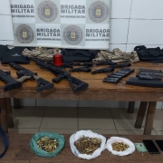 Imagem mostra uma mesa de madeira com muitas armas em cima. Por trás, o banner da Brigada Militar. 