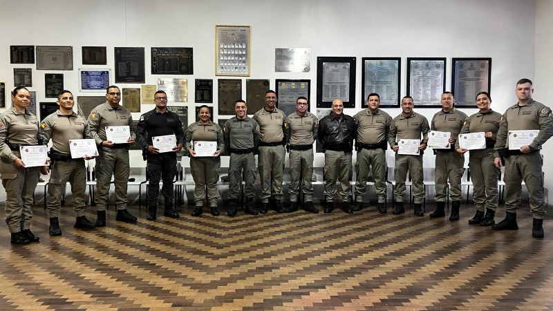 Imagem mostra formandos ostentando os seus certificados em uma sala. Na parede ao fundo, várias placas de formaturas