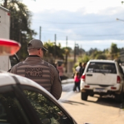 Foto mostra uma viatura parada com o giroflex ligado em uma avenida. Mais à frente, movimentação de pessoas e um carro branco parado.