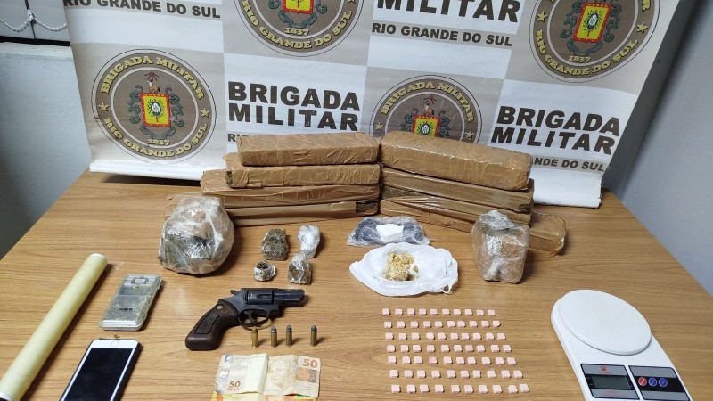 Foto mostra porções de drogas empilhadas em cima de uma mesa, junto a balanças de precisão, celular, notas de dinheiro e uma arma. Em segundo plano, o banner da Brigada Militar.