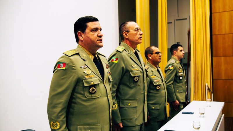 Foto mostra quatro oficiais da BM perfilados em uma mesa, em posição de respeito, durante a execução de hino.