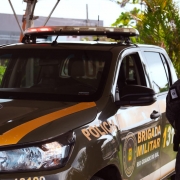 Policial militar do batalhão de choque ao lado de uma viatura verde em posição de respeito.