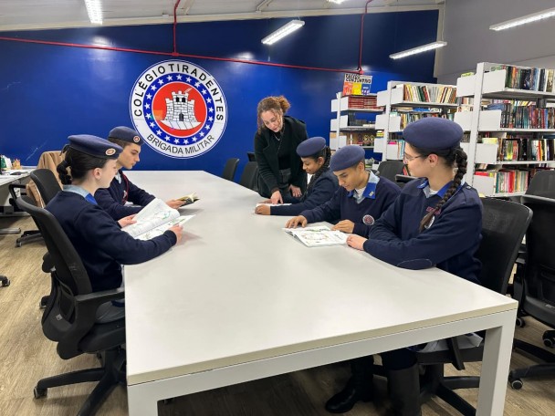 Estudantes aparecem em volta de uma mesa na biblioteca. Estão fardados e ao fundo aparecem as estantes com os livros e uma parede azul com o brasão do CT. 