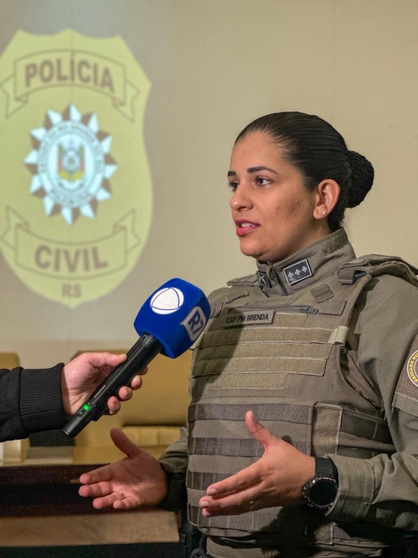 Imagem mostra uma capitã PM da Brigada Militar, fardada, concedendo entrevista para uma equipe de imprensa. Um microfone azul é segurado próximo à policial. Ao fundo, aparece a projeção do brasão da Polícia Civil em uma tela.