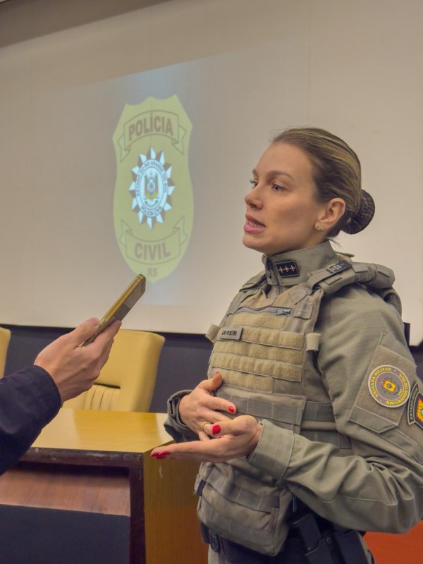 Imagem mostra uma capitã PM da Brigada Militar, fardada, concedendo entrevista em um auditório. Uma pessoa segura um celular próximo à policial para gravar a fala. Ao fundo, há uma mesa, cadeiras e a projeção do brasão da Polícia Civil em uma tela.