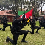 Foto mostra policiais militares ajoelhados em um gramado, com armas apontadas para cima, com a bandeira do RS em fundo