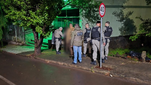 PMs aparecem na cal&ccedil;ada do terreno ao lado da casa onde um agressor manteve tr&ecirc;s pessoas da fam&iacute;lia em c&aacute;rcere privado por sete horas. Imagem noturna com asfalto molhado e luz verde da frente da resid&ecirc;ncia. 