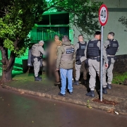 PMs aparecem na cal&ccedil;ada do terreno ao lado da casa onde um agressor manteve tr&ecirc;s pessoas da fam&iacute;lia em c&aacute;rcere privado por sete horas. Imagem noturna com asfalto molhado e luz verde da frente da resid&ecirc;ncia. 